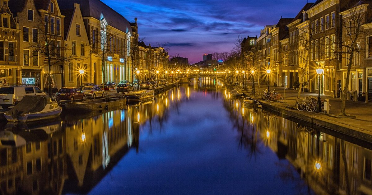 Nederlandse gracht bij avondschemering, met historische gevels, boten en weerspiegelende lantaarns.